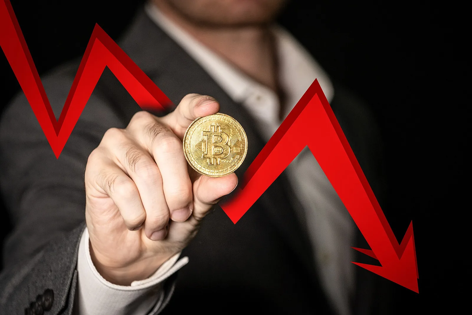 Concept photo of a hand holding a bitcoin token in front of a falling red arrow, illustrating the crypto price fall after the Binance hack